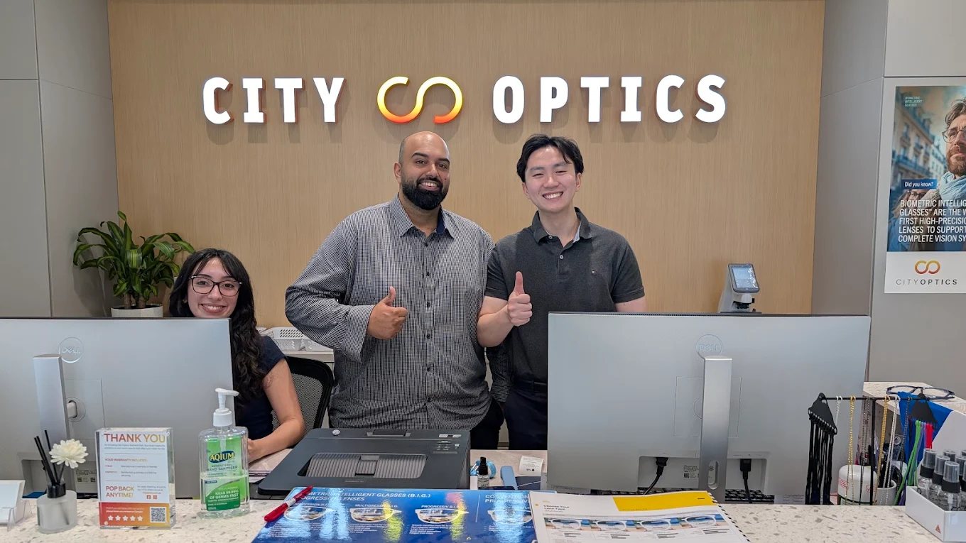 The City Optics team smiling and giving a thumbs up at their front counter under the City Optics sign